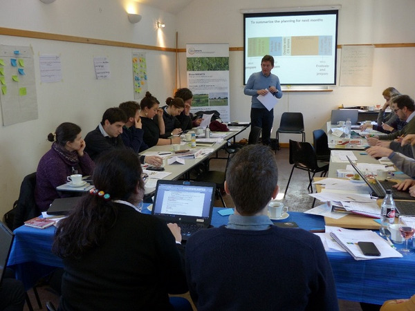 A person standing in front of a group of people at tables, with a presentation in the background